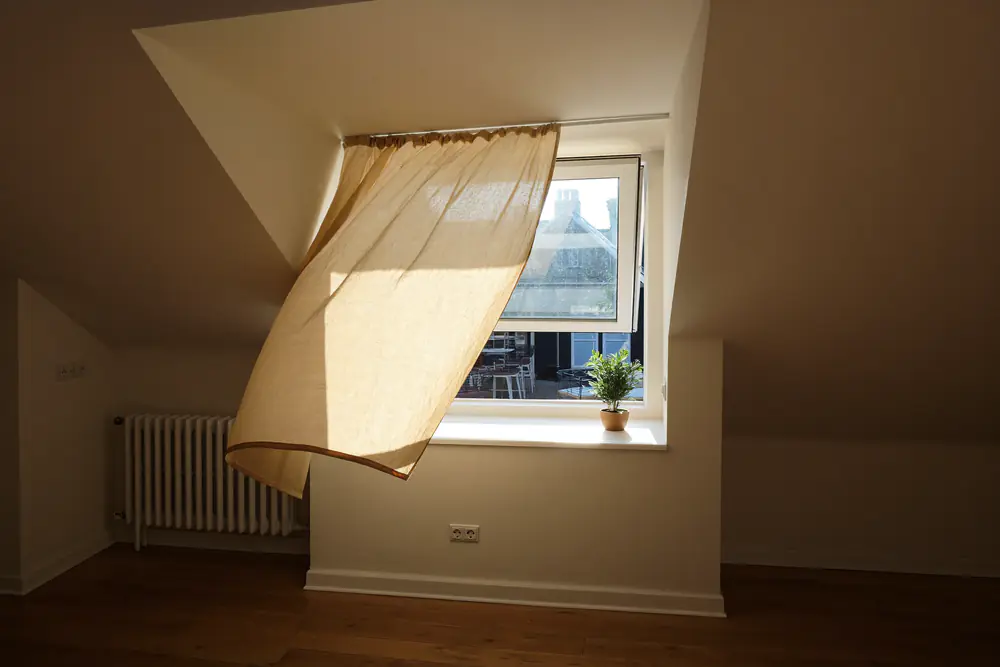 Binnenkant van een dakkapel met een openstaand draaikiepraam en een gordijn dat in de wind beweegt