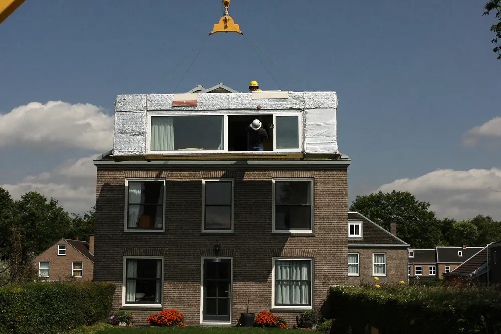 Een kraan tilt een in folie verpakte prefab dakkapel op het dak van een bakstenen woning