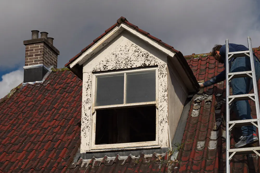 Verweerde houten dakkapel met afbladderende verf, een man op een ladder inspecteert de schade