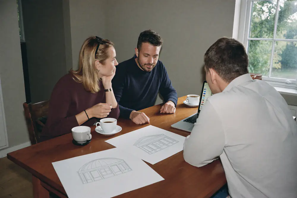 Twee bewoners en een adviseur bekijken tekeningen van een dakkapel aan een keukentafel