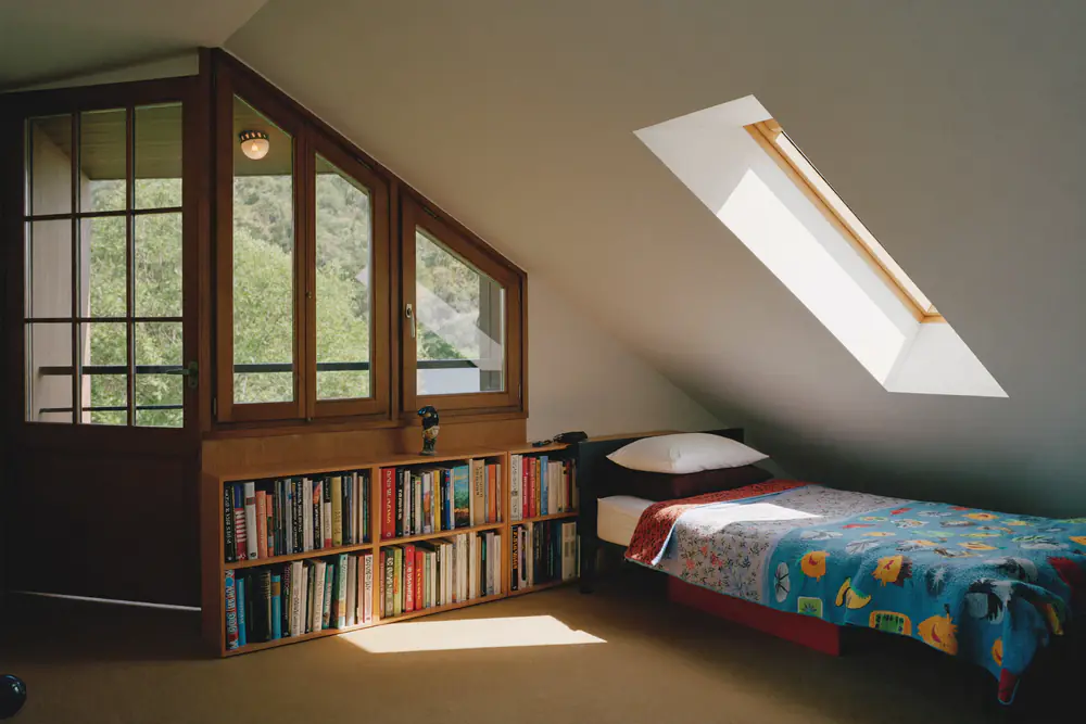 Kinderkamer op zolder met een eenpersoonsbed met kleurige deken, een boekenkast en grote ramen