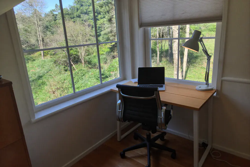 Houten bureau met laptop en bureaulamp voor een groot raam met uitzicht op bomen